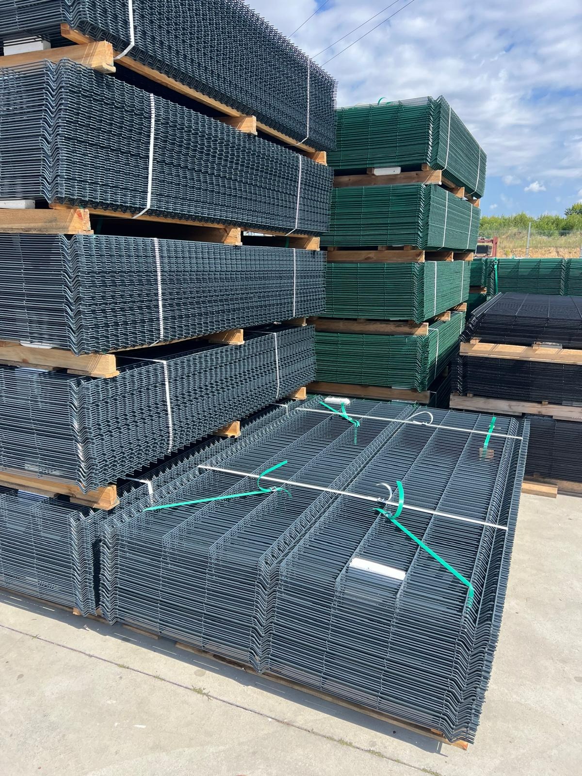 Stosy paneli ogrodzeniowych w kolorze grafitowym i zielonym, zabezpieczone taśmami na drewnianych paletach, przygotowane do transportu, w tle widoczne niebo i zieleń.