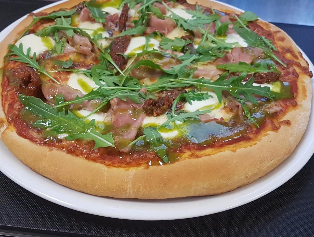 Pizza z szynką parmeńską, mozzarellą, suszonymi pomidorami i rukolą, polana zielonym pesto, serwowana na białym talerzu.