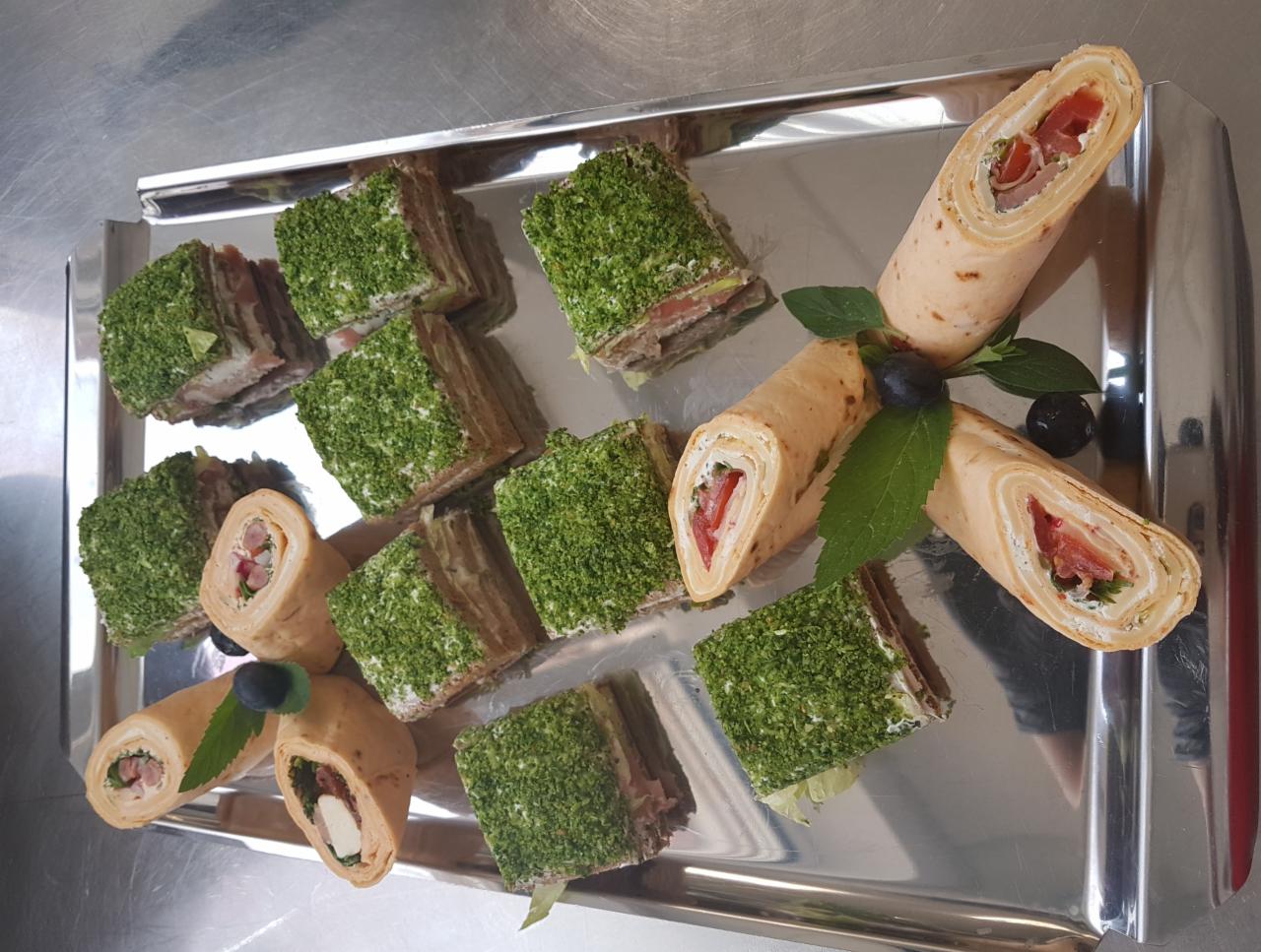 Srebrna taca z roladkami z tortilli z nadzieniem i kanapkami pokrytymi zieloną posypką, udekorowana listkami mięty i borówkami.