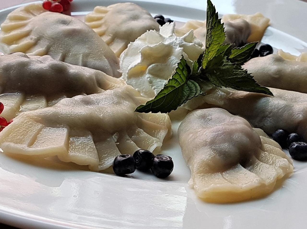 Talerz z pierogami z owocami, udekorowanymi bitą śmietaną, borówkami i listkami mięty, z dodatkiem czerwonych porzeczek.