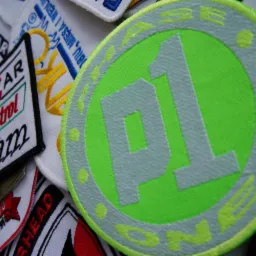 Kolekcja naszywek z haftowanymi logo i napisami, w tym okrągła, neonowo-zielona naszywka z szarym napisem 'P1' i obwódką z żółtymi detalami, ułożona obok naszywki 'Rycar Castrol Team'.