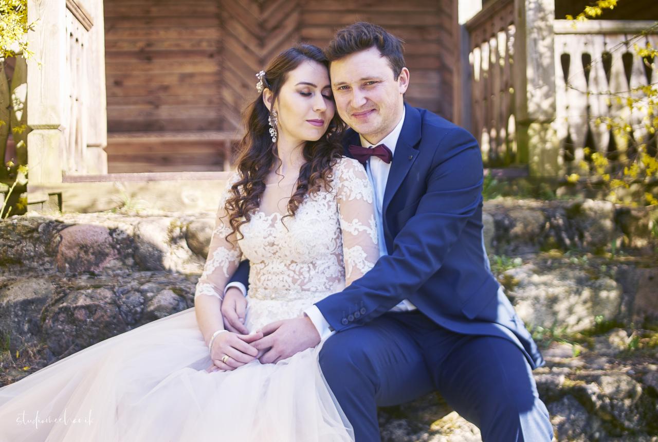 Para młoda siedzi na kamiennych schodach przed drewnianym budynkiem, panna młoda w koronkowej sukni z długim rękawem, pan młody w granatowym garniturze z bordową muchą.