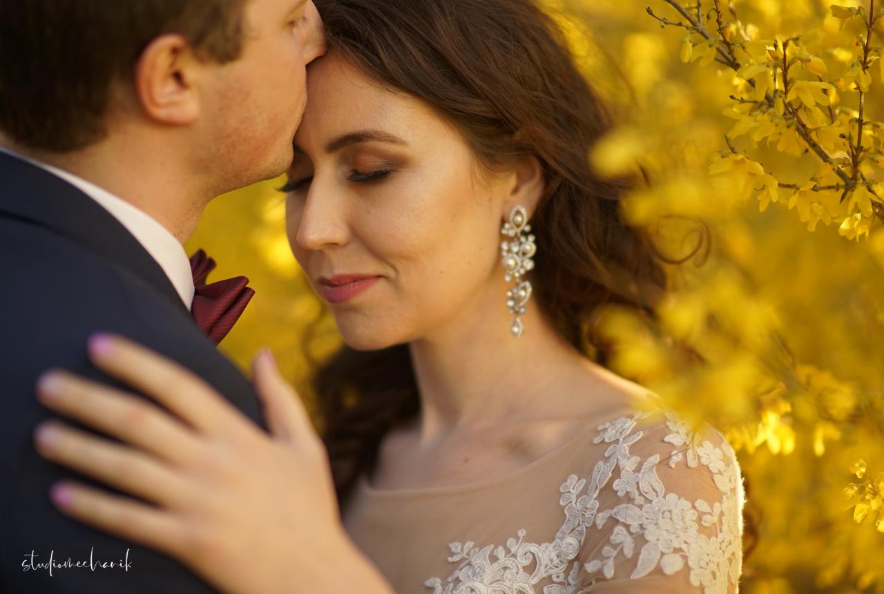 Panna młoda z zamkniętymi oczami, w sukni z koronkowym zdobieniem, otoczona kwitnącym forsycją, mężczyzna w granatowym garniturze całuje ją w czoło.