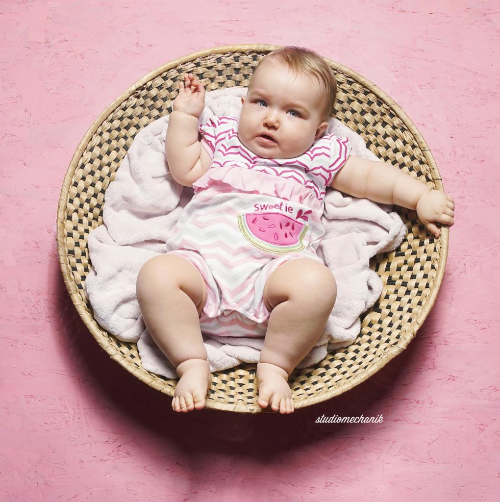Niemowlę w paski w koszyku na różowym tle, ubrane w rampers z aplikacją arbuza.
