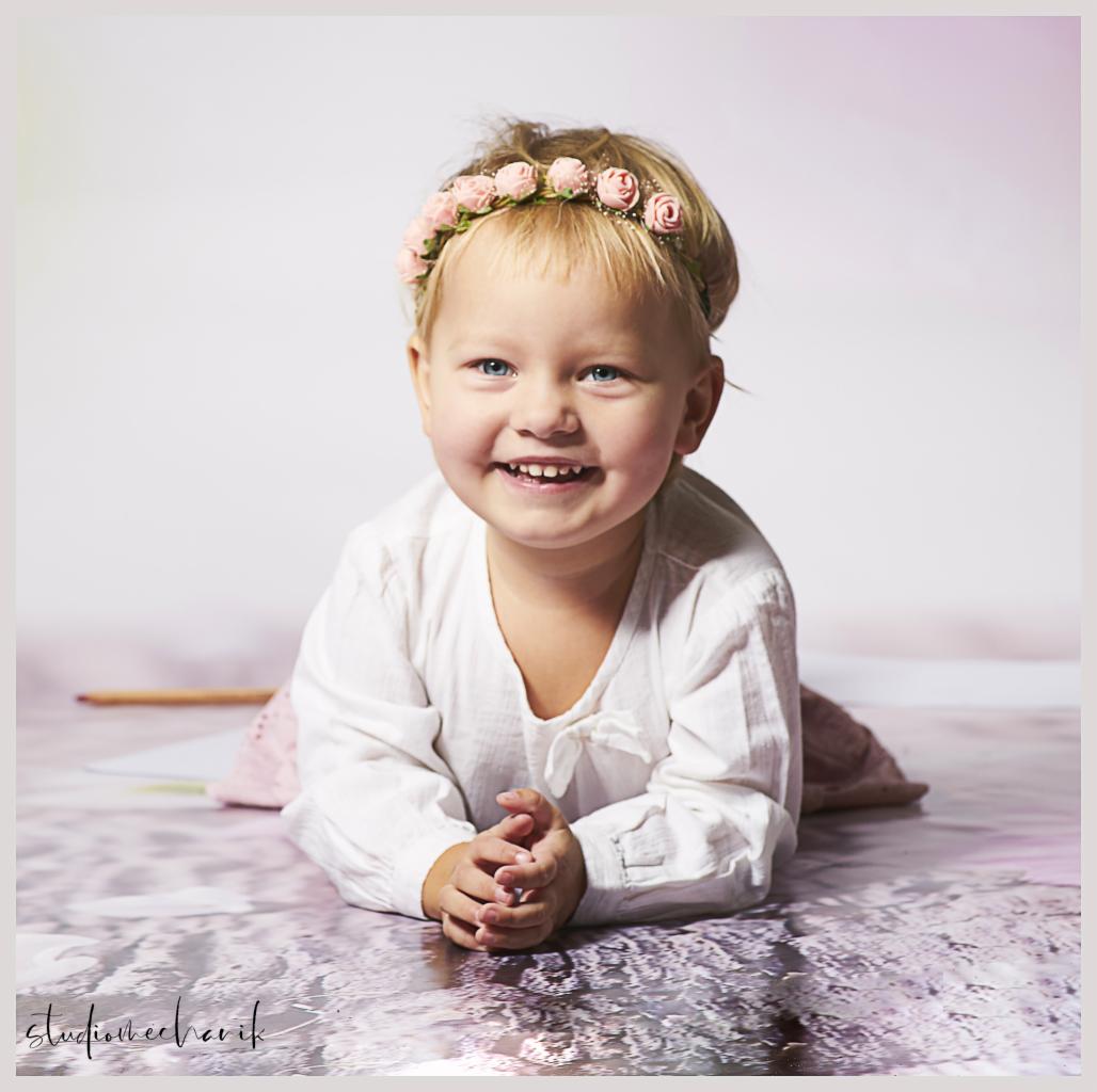 Uśmiechnięta dziewczynka z blond włosami i wiankiem z różowych różyczek leży na brzuchu na wzorzystym tle, dłonie złożone przed sobą, w tle widoczny ołówek i kartka.