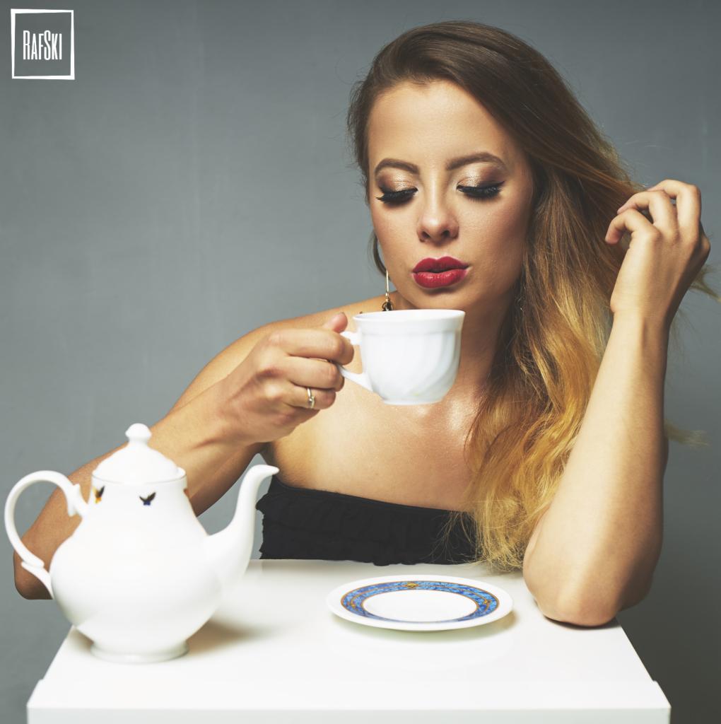 Portret kobiety z blond ombre pijącej z białej filiżanki, obok biały imbryk z motywem motyli i talerzyk z niebieskim wzorem, neutralne szare tło