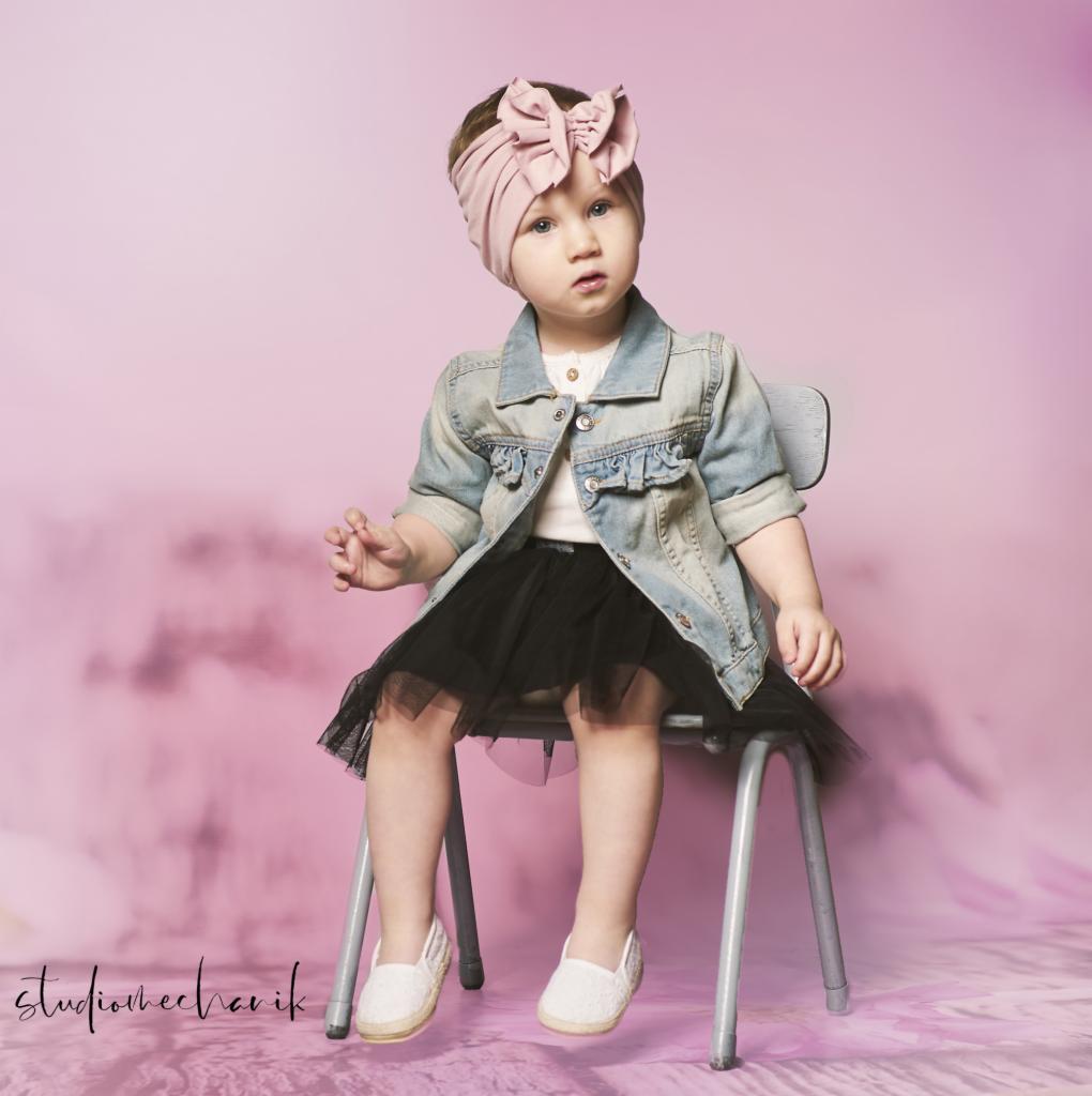 Dziewczynka w dżinsowej kurtce i tiulowej spódniczce siedzi na metalowym krześle na tle różowego, abstrakcyjnego tła.