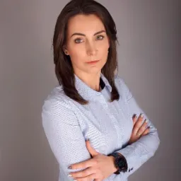 Portret kobiety w błękitnej koszuli z zegarkiem na ręce, skrzyżowane ramiona, neutralne tło.