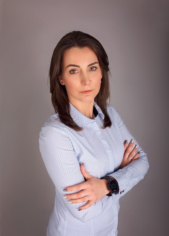 Portret kobiety w błękitnej koszuli z zegarkiem na ręce, skrzyżowane ramiona, neutralne tło.