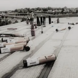 Krycie dachu płaskiego papą termozgrzewalną marki Bauder, widoczne rolki papy, butle gazowe, kominy wentylacyjne i dekarze pracujący na dachu w oddali, widok na miasto w tle.