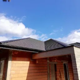 Nowo wybudowany dom z ciemną dachówką i zamontowanymi, czarnymi rynnami, na tle błękitnego nieba z chmurami.