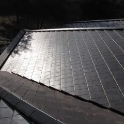 Nowoczesny dach pokryty ciemnymi, prostokątnymi płytkami z metalu, odbijającymi światło słoneczne, z widoczną obróbką blacharską i elementami konstrukcyjnymi.