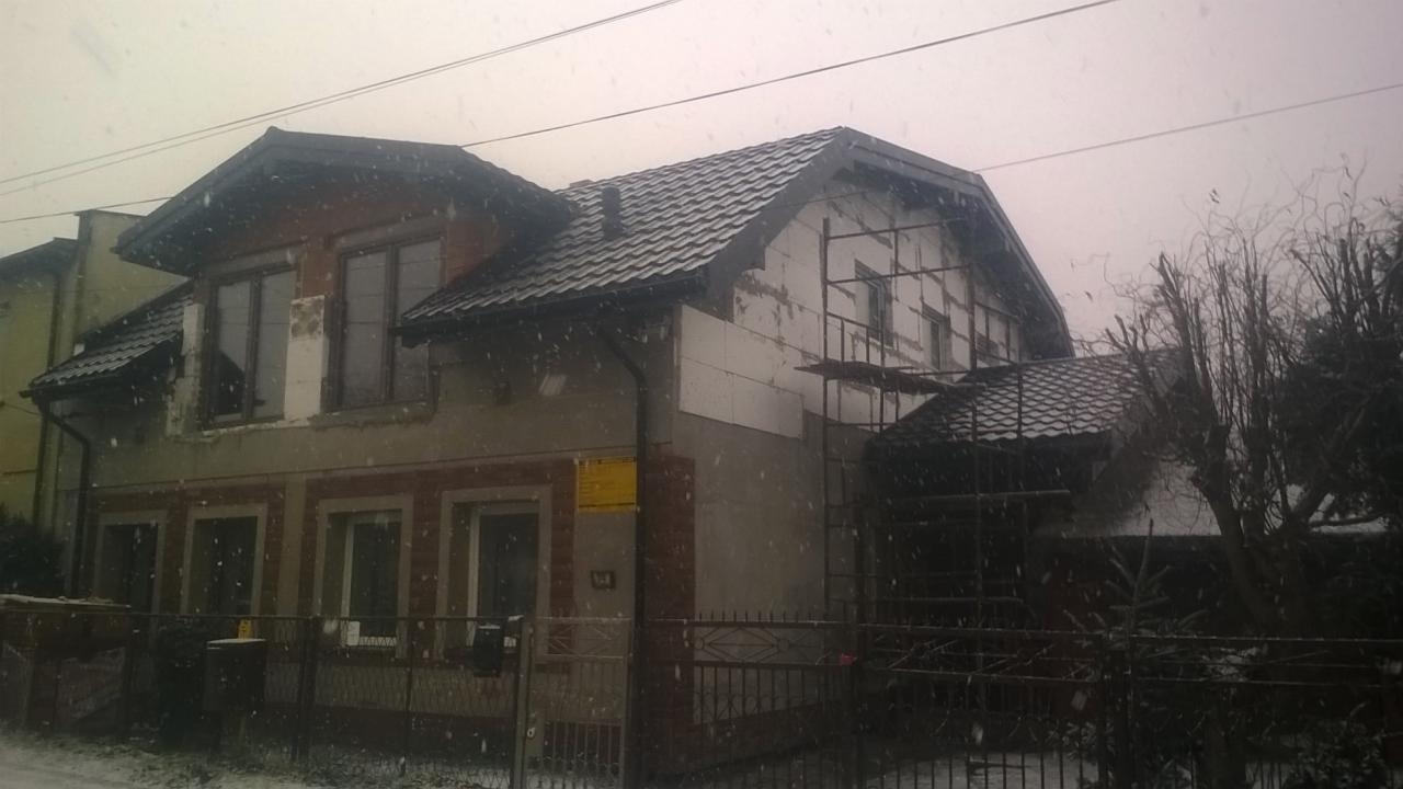 Dom w trakcie ocieplania elewacji i wymiany pokrycia dachowego, widoczne rusztowania i styropian na ścianach, nowy dach z dachówki ceramicznej, zimowa sceneria.