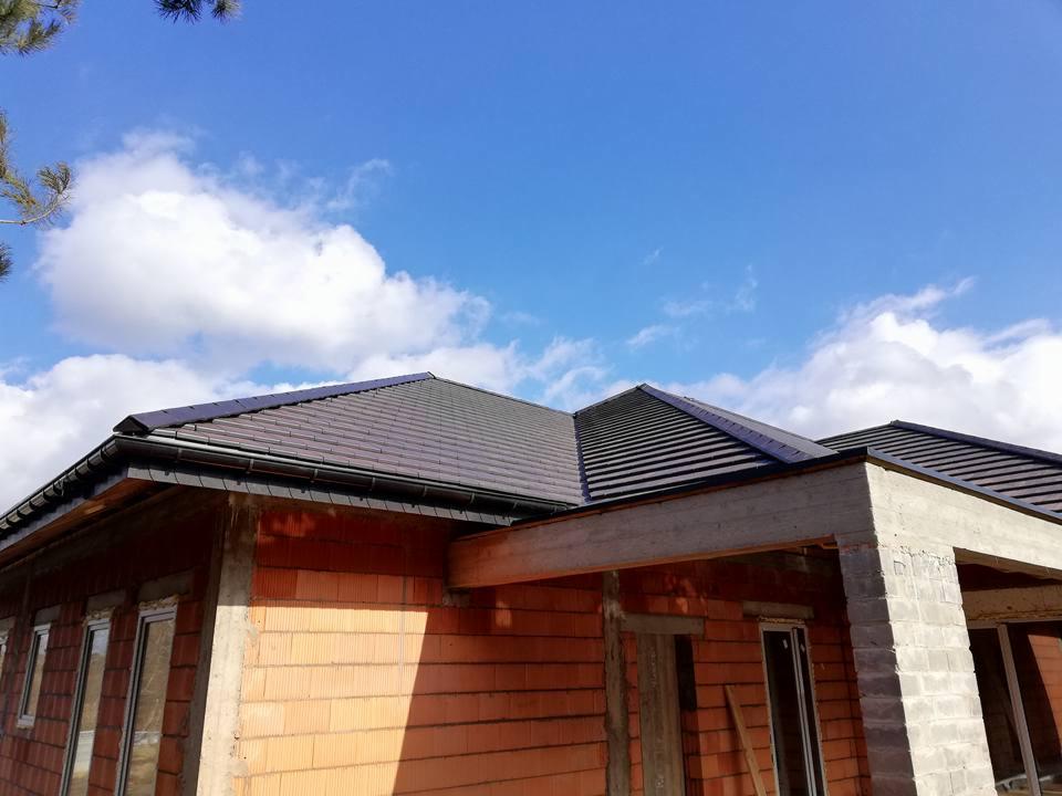 Nowo wybudowany dom z ciemną dachówką i zamontowanymi, czarnymi rynnami, na tle błękitnego nieba z chmurami.