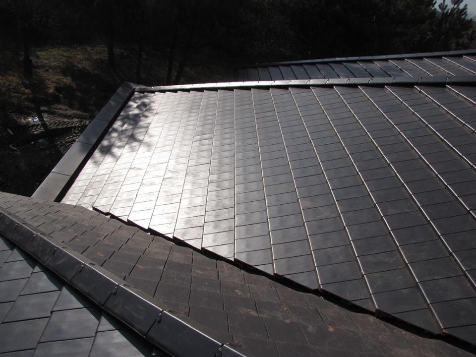 Nowoczesny dach pokryty ciemnymi, prostokątnymi płytkami z metalu, odbijającymi światło słoneczne, z widoczną obróbką blacharską i elementami konstrukcyjnymi.