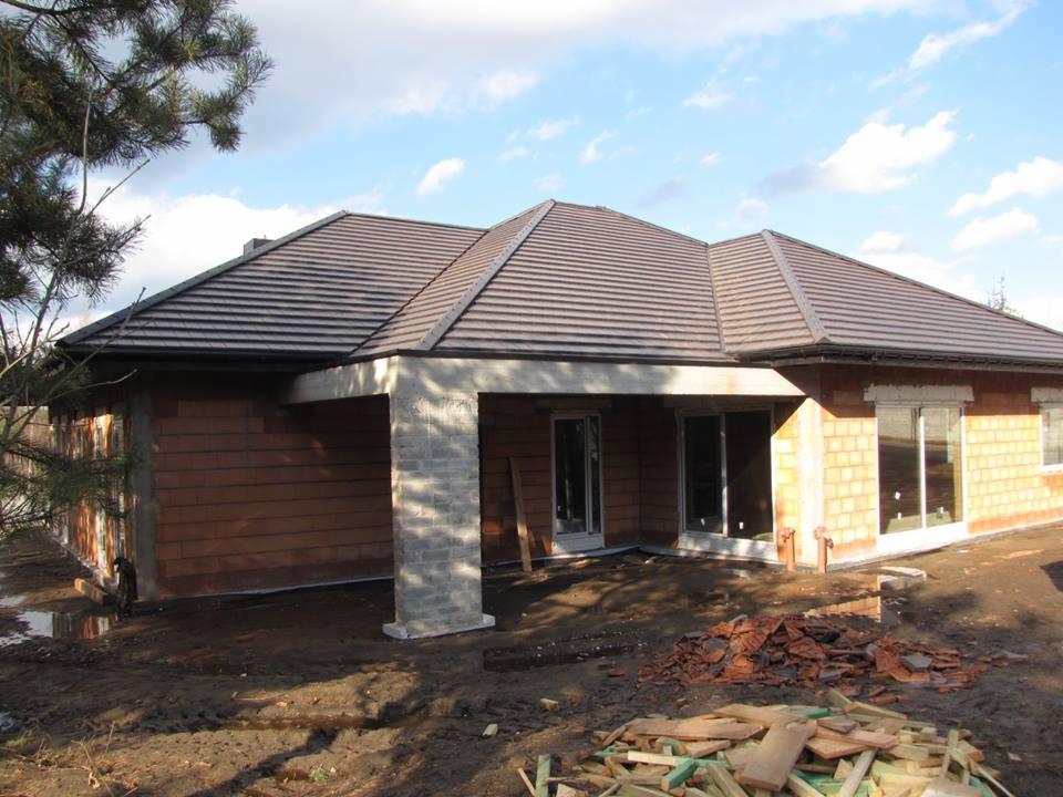 Dom jednorodzinny w trakcie budowy, z częściowo ukończonym, brązowym dachem z dachówki ceramicznej i surowymi ścianami z czerwonej cegły, widoczne ślady prac budowlanych na podwórku.