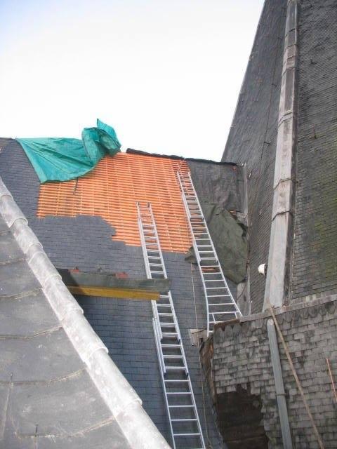 Renowacja dachu kamienicy: fragment dachu z odsłoniętą drewnianą konstrukcją, zabezpieczony plandeką, dwie drabiny opierają się o dach i ścianę budynku.