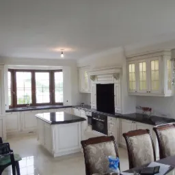 Jasna kuchnia z wyspą i czarnym blatem, widoczna z perspektywy jadalni z kryształowym żyrandolem