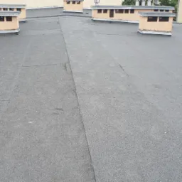 Szary, płaski dach pokryty papą bitumiczną z widocznymi łączeniami, kominy wentylacyjne w tle, budynki mieszkalne w oddali.