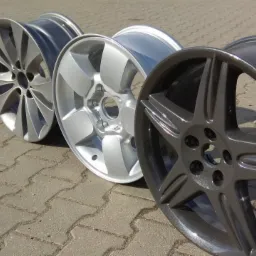 Trzy odnowione felgi aluminiowe w różnych odcieniach szarości, leżące na kostce brukowej, prezentujące różne wzory szprych.