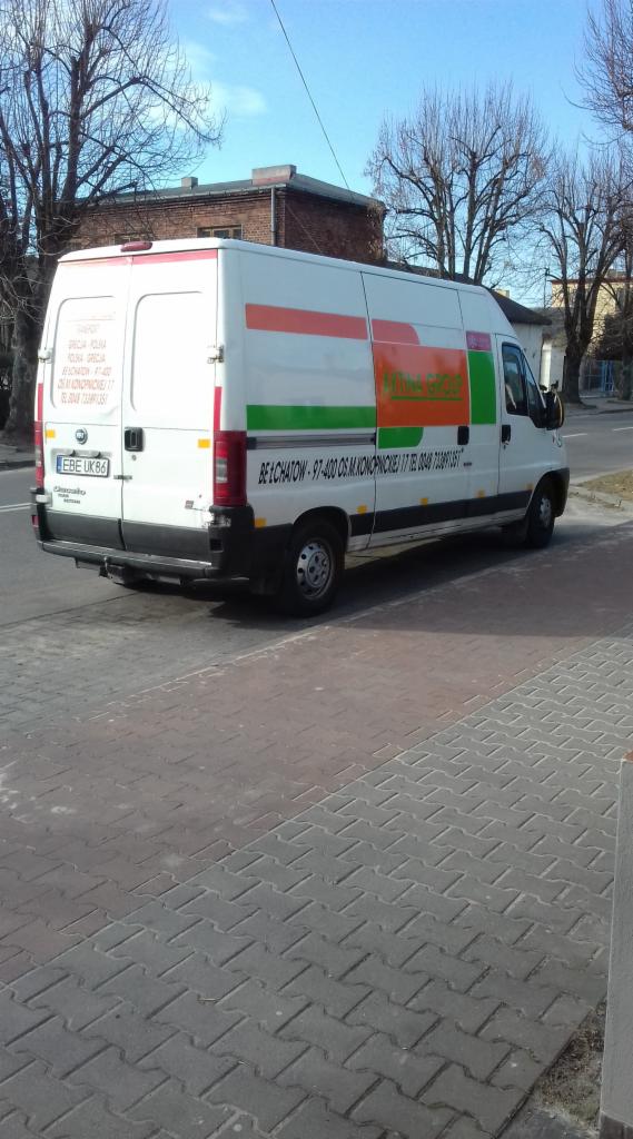 Biały samochód dostawczy firmy transportowej z pomarańczowo-zielonym logo na boku, zaparkowany na chodniku z kostki brukowej w Bełchatowie, widok z tyłu i boku.