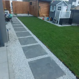 Nowoczesny ogród z ścieżką z kwadratowych płyt betonowych otoczonych białym grysem, prowadzącą obok świeżo założonego trawnika i drewnianej zabudowy z domkiem dla dzieci w tle.