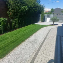 Starannie utrzymany ogród z równo przystrzyżonym trawnikiem, ścieżką z białego żwiru i ozdobnymi drzewami iglastymi wzdłuż ogrodzenia.