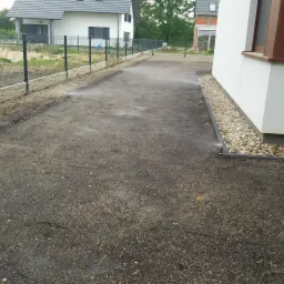 Przygotowany teren pod trawnik z widocznym systemem nawadniania wzdłuż ściany domu i ogrodzeniem panelowym z podmurówką.