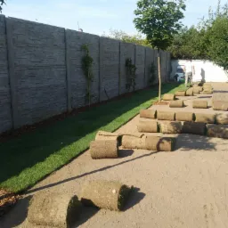 Rozwinięta trawa z rolki na piaszczystym podłożu wzdłuż betonowego ogrodzenia, widoczna osoba w tle.