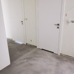 FIRMA KOBO SŁAWOMIR SWĘDRAK - Szara, surowa posadzka z mikrocementu w nowym wnętrzu z dwiema białymi drzwiami. Widoczne niedokończone wykończenia ścian i sufitu. Minimalistyczny design.