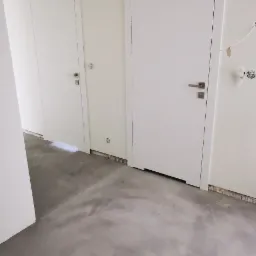 Szara, surowa posadzka z mikrocementu w nowym wnętrzu z dwiema białymi drzwiami. Widoczne niedokończone wykończenia ścian i sufitu. Minimalistyczny design.