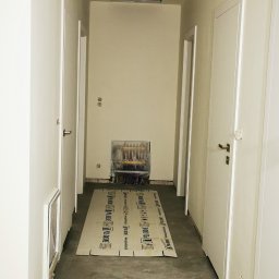 FIRMA KOBO SŁAWOMIR SWĘDRAK - Korytarz w trakcie remontu: zabezpieczone rury grzewcze, podłoga betonowa, kartony ochronne. Widoczne białe drzwi i sufit z otworem.