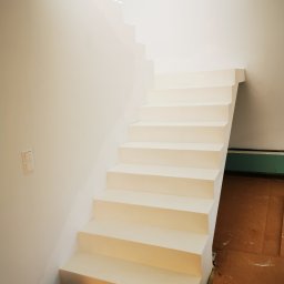 FIRMA KOBO SŁAWOMIR SWĘDRAK - Białe, minimalistyczne schody w jasnym wnętrzu, widok z dołu. Świeżo pomalowane, z widocznymi detalami wykończenia i zabezpieczeniem podłogi kartonem.