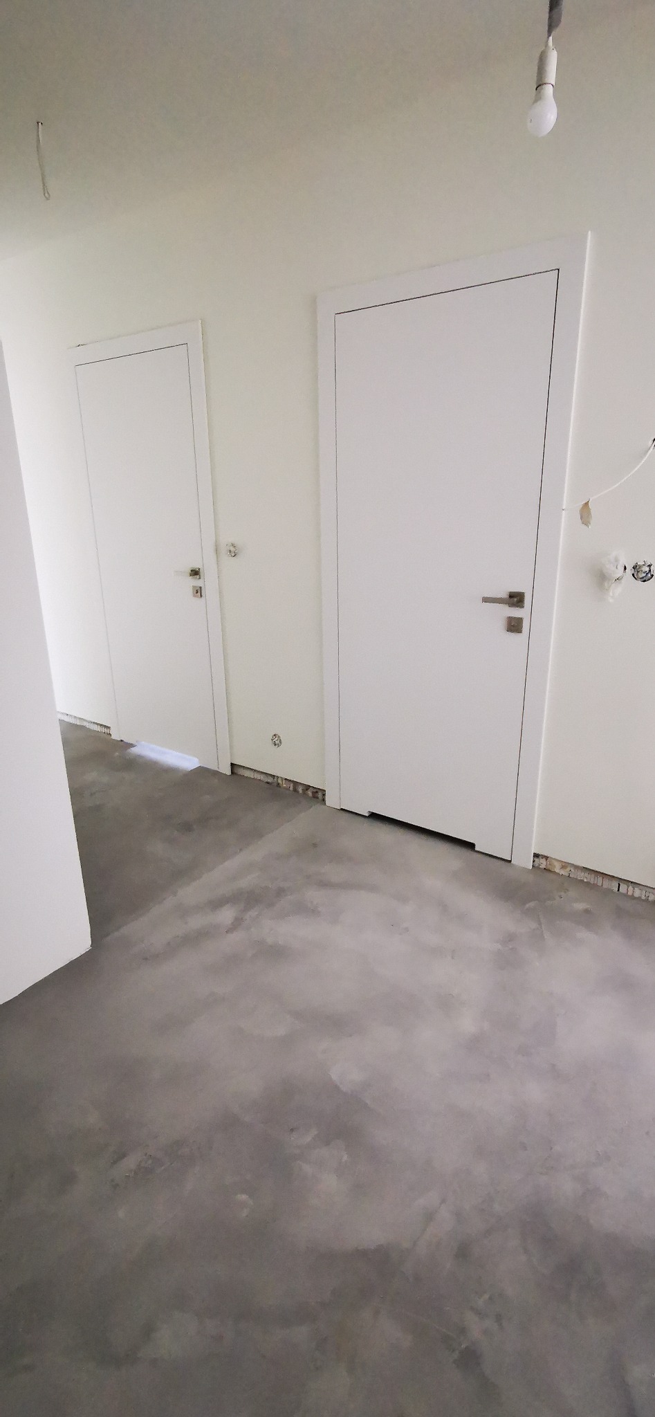 Szara, surowa posadzka z mikrocementu w nowym wnętrzu z dwiema białymi drzwiami. Widoczne niedokończone wykończenia ścian i sufitu. Minimalistyczny design.