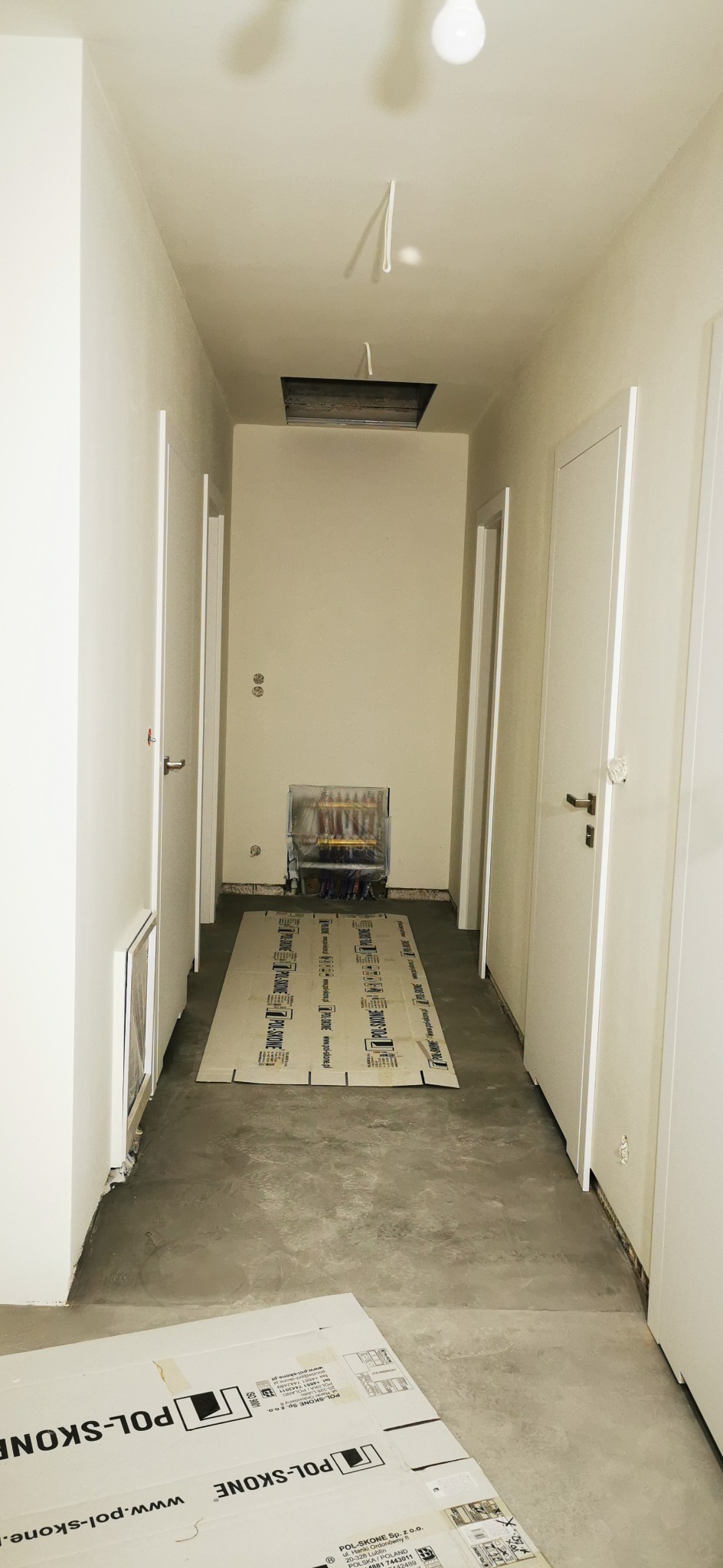 Korytarz w trakcie remontu: zabezpieczone rury grzewcze, podłoga betonowa, kartony ochronne. Widoczne białe drzwi i sufit z otworem.