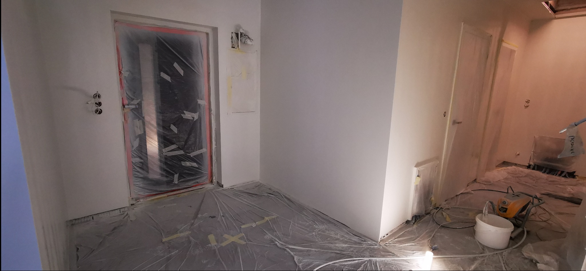 Przygotowane do malowania wnętrze: zabezpieczone folią podłogi i drzwi, świeżo pomalowane ściany na biało, widoczne puszki elektryczne i sprzęt malarski.