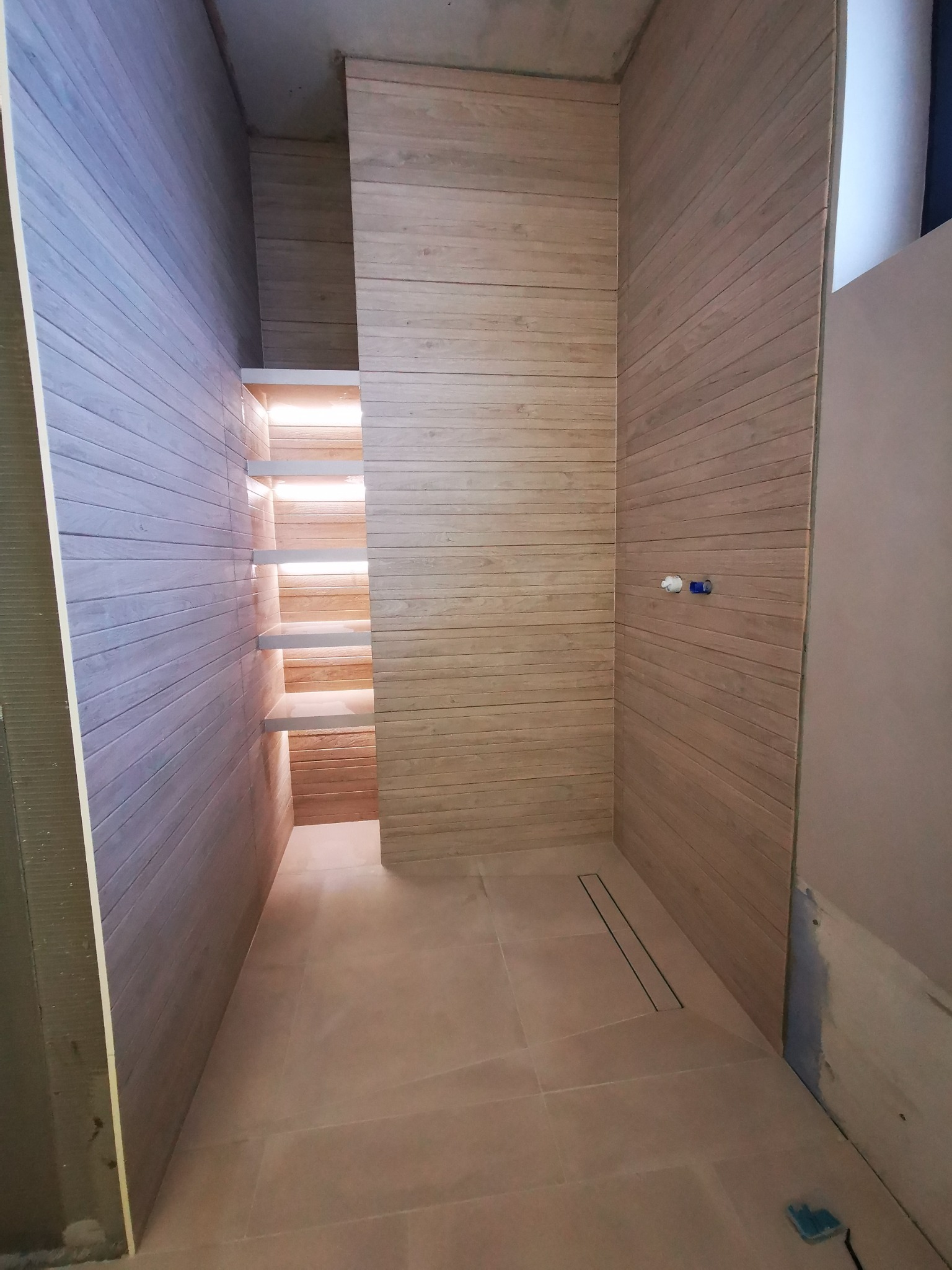 Wykończona łazienka z drewnopodobnymi płytkami na ścianach i podłodze, widoczny odpływ liniowy i półki wnękowe z podświetleniem. Częściowo wykończone ściany.