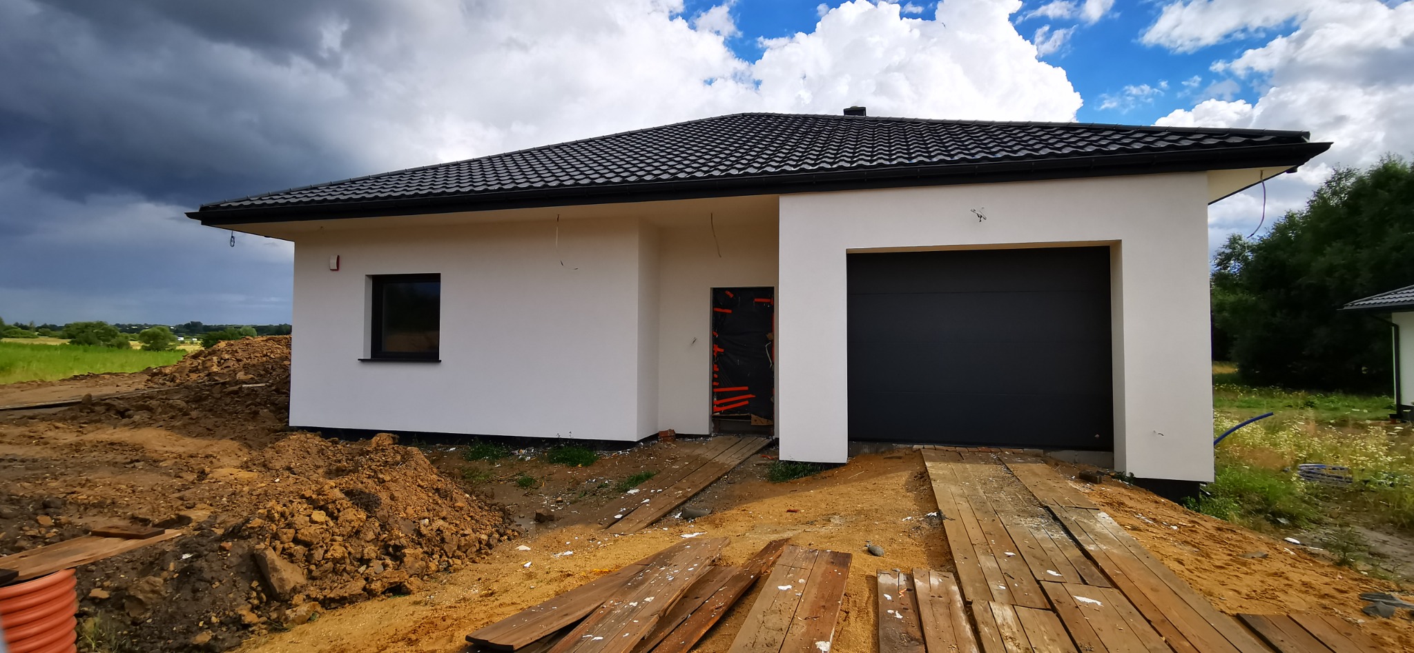Nowoczesny, parterowy dom z garażem w trakcie budowy. Biała elewacja, czarny dach i brama garażowa. Widoczne prace ziemne i przygotowanie terenu.