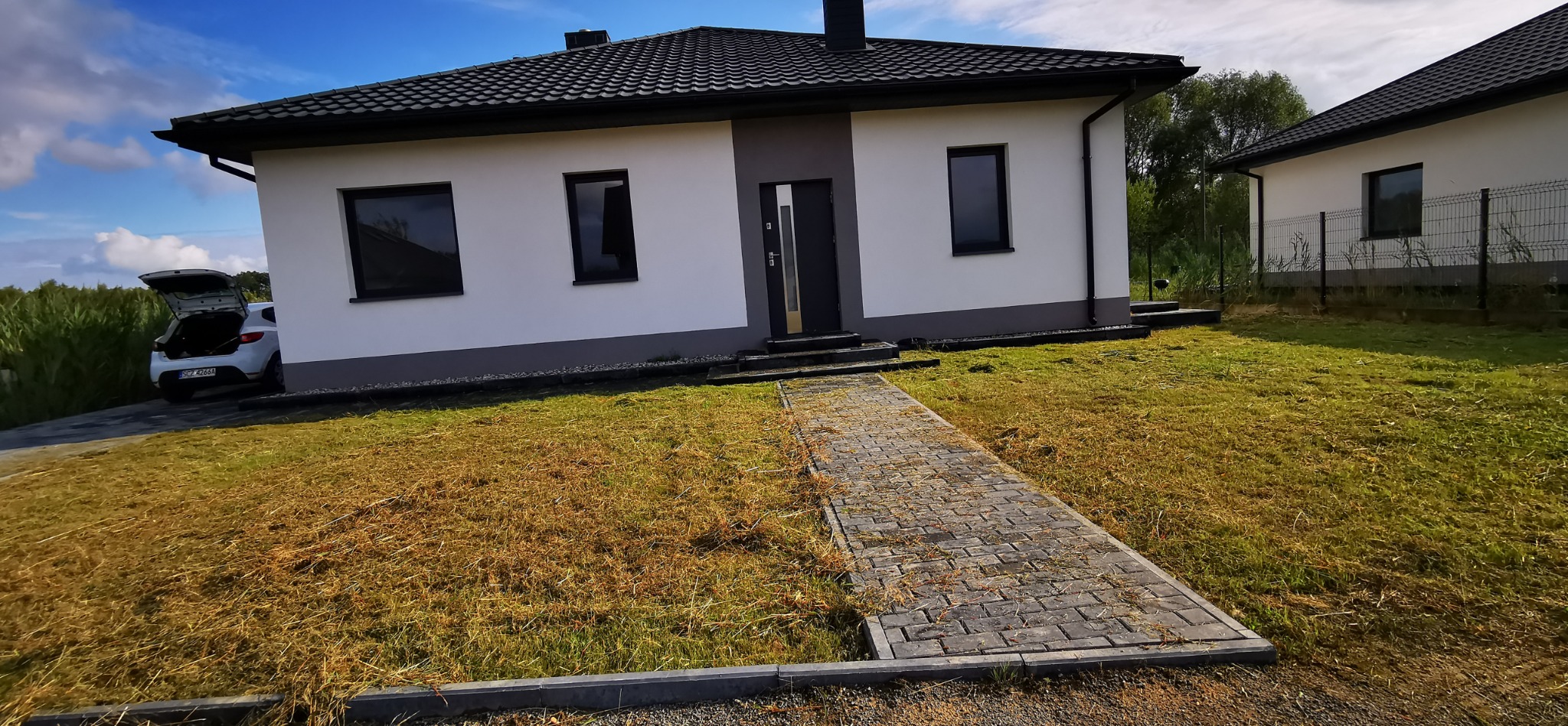 Nowoczesny, parterowy dom z szarą elewacją i czarnymi oknami, widziany z perspektywy chodnika z kostki brukowej. Na trawniku resztki skoszonej trawy. Auto z otwartym bagażnikiem.