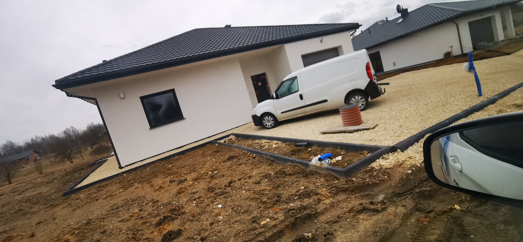 Nowoczesny dom jednorodzinny z ciemną dachówką, jasną elewacją i podjazdem z kamienia. Widoczny samochód dostawczy i przygotowany teren pod ogród.