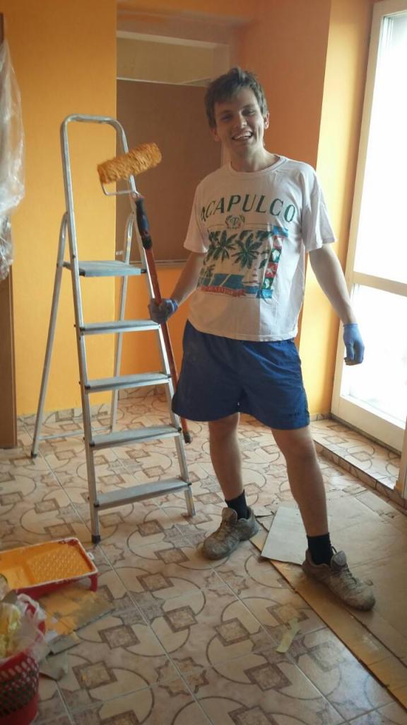 Uśmiechnięty mężczyzna w niebieskich spodenkach i koszulce Acapulco, trzymający wałek malarski na tle pomarańczowej ściany, drabiny i podłogi wyłożonej kafelkami w trakcie malowania mieszkania.