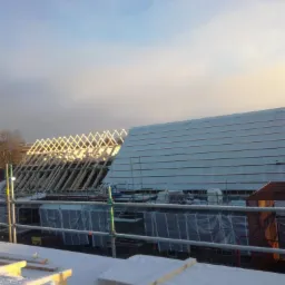 Konstrukcja drewniana dachu w trakcie budowy, częściowo przykryta płytami, lekko ośnieżona, widoczne rusztowania i zabezpieczenia.