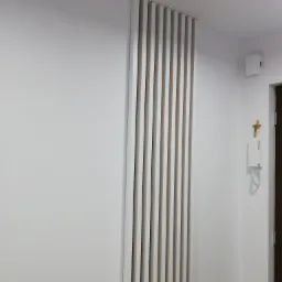 Biała ściana z dekoracyjnymi, pionowymi panelami z listew, obok domofon i krzyż na ścianie w jasnym wnętrzu.