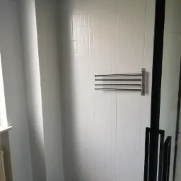 Wykończona łazienka z białymi, kwadratowymi płytkami na ścianach, czarnym sufitem i podłogą imitującą marmur. Widoczny grzejnik drabinkowy na ścianie oraz fragment czarnej kabiny prysznicowej.