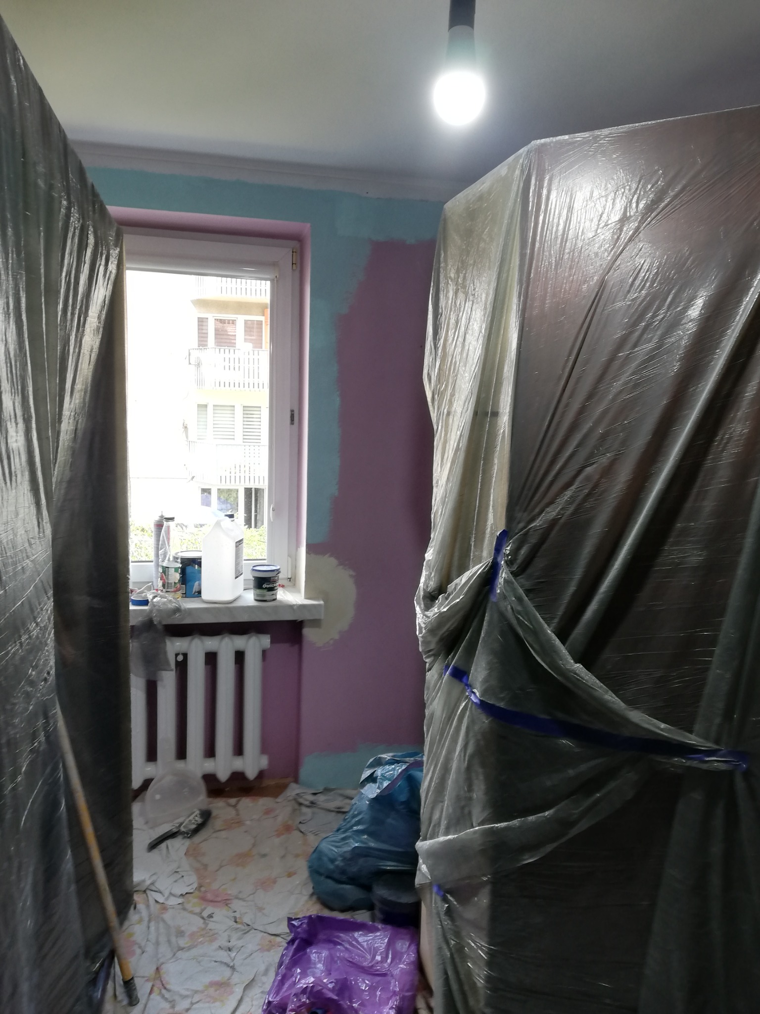 Pokój w trakcie malowania: zabezpieczone meble folią, ściany częściowo pomalowane na różowo i niebiesko, widoczne puszki z farbą na parapecie okiennym, podłoga zabezpieczona folią malarską.