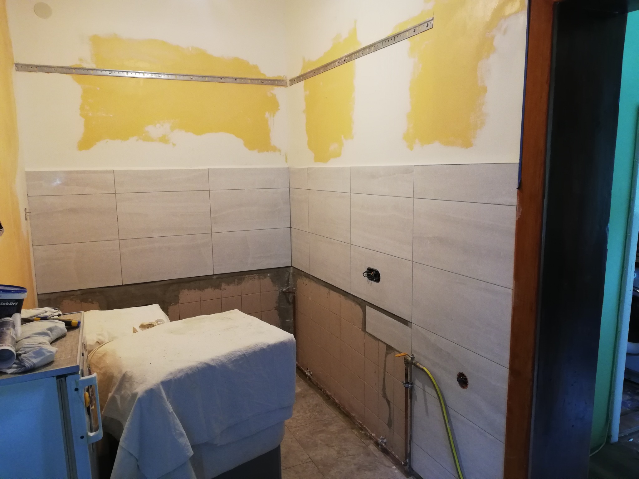 Łazienka w trakcie remontu: częściowo położone płytki ścienne, fragmenty ścian z żółtą farbą, metalowe szyny montażowe pod sufitem, zakryte meble, widoczne rury instalacji wodnej.