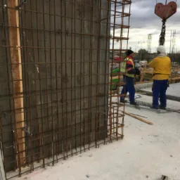 Konstrukcja zbrojeniowa szalunku betonowego z widocznymi pracownikami w tle i żurawiem na budowie.