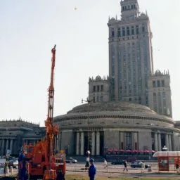 Wiertnica w kolorze pomarańczowym na tle Pałacu Kultury i Nauki w Warszawie, widoczni pracownicy w niebieskich kombinezonach, rury wiertnicze na trawie.