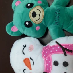 Dwa pluszowe, miękkie zabawki: turkusowy miś z haftowanym imieniem 'Wiktor' i biały bałwanek ze szkarłatnym szalikiem w kropki, leżące na ciemnym blacie.