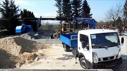 Biała ciężarówka z niebieską skrzynią ładunkową na placu budowy. W tle widoczny podnośnik z ramieniem, koparka i hałdy piasku oraz gruzu.